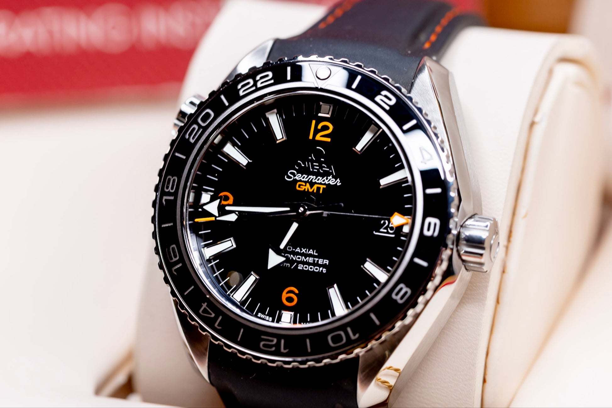 2024 Omega Seamaster Planet Ocean GMT watch displayed on a soft cushion with black strap and vibrant orange accents.